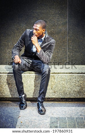 Young black man thinking outside. Dressing in a blazer, pants, leather shoes, a hand touching his chin, a young black guy is sitting on a marble stone bench, into deeply thinking. Street Fashion.