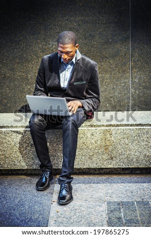 Young black man working outside. Dressing in a blazer, pants, leather shoes, a young black guy is sitting on a marble stone bench, looking down, working on a laptop computer.
