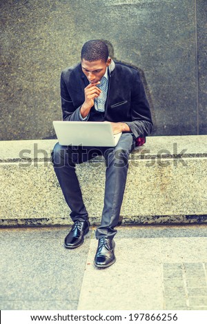 Young black man working outside. Dressing in a blazer, pants, leather shoes, a young black guy is sitting on a marble stone bench, looking down, a hand touching his chin, working on a laptop computer.