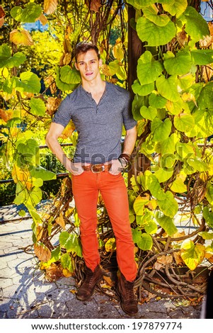Man casual fashion.Dressing in a gray long sleeves with roll-tab Henley shirt, red jeans, brown boot shoes, a young handsome guy is standing by green plants with big leaves, charmingly looking at you.
