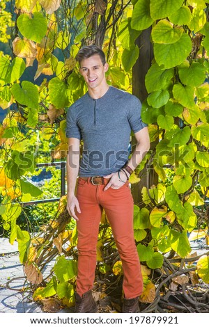 Man casual fashion.Dressing in a gray long sleeves with roll-tab Henley shirt, red jeans,  a young handsome guy is standing by green plants with big leaves, smilingly looking at you.