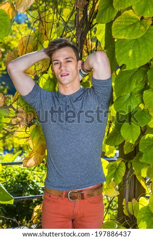Man Casual Fashion. Dressing in a gray long sleeves with roll-tab Henley shirt, red jeans, hands putting in the back of head, a young handsome guy is standing by green plants, looking at you.