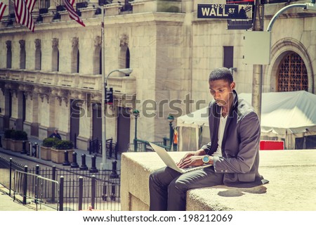 Young man working on street. A young black college student is sitting outside, working on a laptop computer, looking down, thinking.  Wall Street sign in the background.