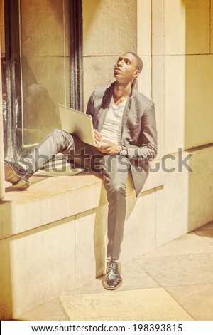 Young Man Working on Computer. Wearing a white under wear, fashionable jacket, pants, shoes, a computer on lap, a young black college student is sitting against a window frame, looking up, thinking.