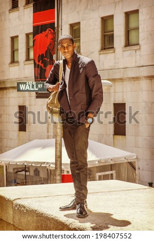 Street Fashion. Wearing a white under wear, fashionable jacket, pants, leather shoes, carrying a shoulder bag,  a young black college student is standing on Wall Street, hopefully looking at you.