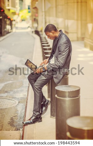 Young man working on street. A young black college student is sitting outside an office building, typing on a laptop computer, reading, thinking.