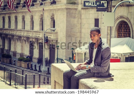 Young man working on street. A young black college student is sitting outside, typing on a laptop computer, looking down, thinking.  Wall Street sign in the background.