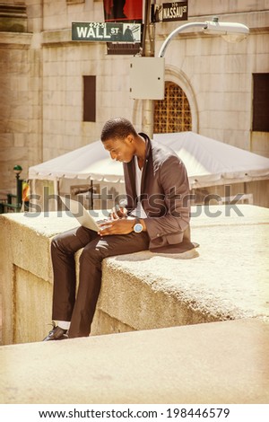 Young man working on street. A young black college guy is sitting outside, looking down, working on a laptop computer. Wall Street sign in the background.