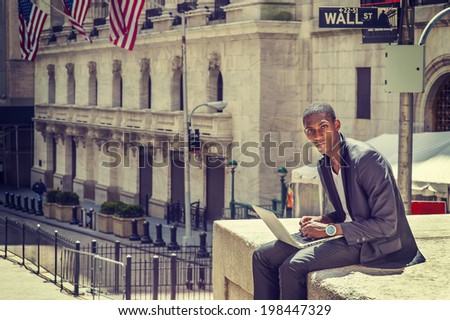 Young man working on street. A young black college student is sitting outside, working on a laptop computer, thinking.  Wall Street sign in the background.