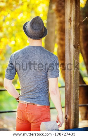 Man Back View. Dressing in a gray long sleeves with roll-tab Henley shirt, wearing a woolen Fedora hat, a young sexy guy is standing, looking back at  golden foliage background in a autumn afternoon.