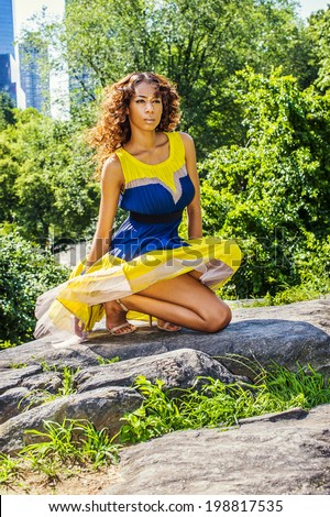 Woman Waiting for You. Dressing in sleeveless, yellow and blue pattern skirt dress, a young pretty black girl with long curly hair is squatting on rocks in the wind, passionately looking forward.