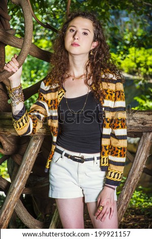 Dressing in black under wear, patterned fashion jacket, short pants, chunky chain bracelet, a teenager girl with curly long hair is standing in wood area, waiting for you. Woman Fashion in Location.