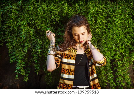 Thinking on green. Dressing in black under wear, patterned fashion jacket, hands holding leaves, a teenager girl with curly long hair is standing by planted wall, looking down, lost in thought.