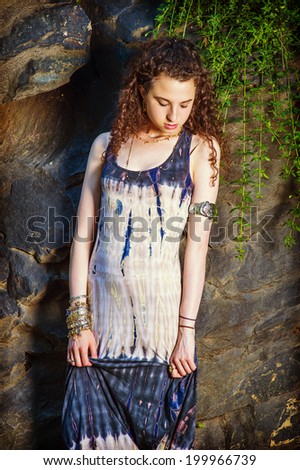 Unhappy Girl. Dressing in patterned long dress, chunky chain bracelet, arm cuff bracelet, a beautiful teenager girl with curly long hair is standing by rocky wall, looking down, sad, thinking.