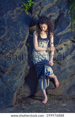 Teen Fashion.Dressing in patterned long dress, chunky chain bracelet, arm cuff bracelet, barefoot, a pretty teenager girl with curly long hair is standing against rocky wall, looking down, thinking.
