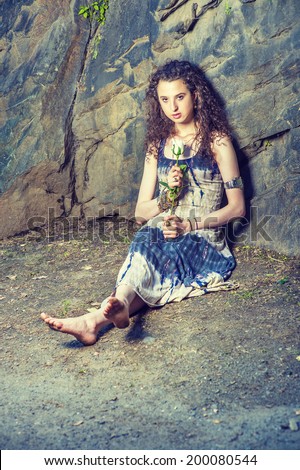 Waiting for You. Dressing in patterned dress, bracelet, barefoot, a pretty teenager girl with curly long hair is sitting on ground against rocky wall, holding a white rose, sincerely looking at you.