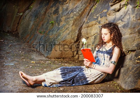 Reading Outside. Dressing in long dress, bracelet, barefoot, a pretty teenager girl with curly long hair is sitting on ground against rocks, holding a red book and a white rose, relaxing, reading.