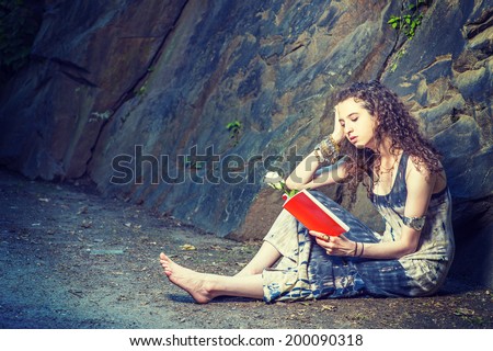 Girl Reading Outside. Wearing long dress, barefoot, a pretty teenage college student with curly long hair is sitting on ground against rocks, holding a red book and a white rose, reading, relaxing.