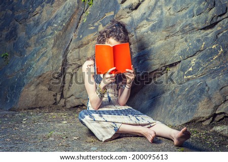Girl Thinking Hard. Wearing long dress, bracelet, barefoot, a pretty teenage college student with curly long hair is sitting on ground against rocks, holding a red book covering her face, thinking.