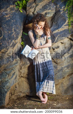 College Student. Dressing in long dress, chunky chain bracelet, arm cuff bracelet, barefoot, a teenage girl with curly long hair is standing against rocks, holding a book, talking on cell phone.