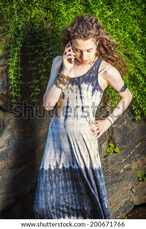 Girl Making Phone Call. A hand holding a cell phone, a hand resting on hips, a teenage college student with curly long hair is standing against rocks, looking down, seriously listening to the phone.