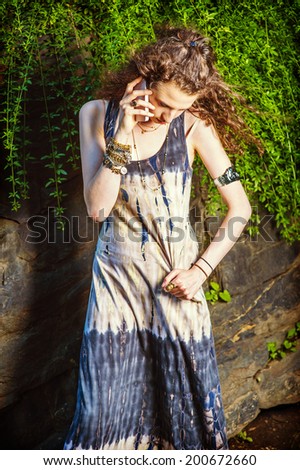 Girl Making Phone Call. A hand holding a cell phone, a hand grasping dress, a teenage college student with curly long hair is standing against rocks, looking down, sad, seriously listening to phone.