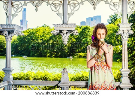 Lady Missing You. Dressing in sleeveless, long dress, a teenage girl is standing inside a pavilion, hands holding a white rose, looking down, smelling, thinking. people rolling boats in background,.