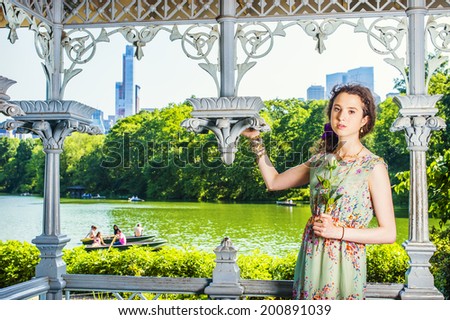 Lady Waiting for You. Dressing in sleeveless, long dress, a pretty teenage girl is standing inside a pavilion at park, hand holding a white rose, looking at you. people rolling boats in background.