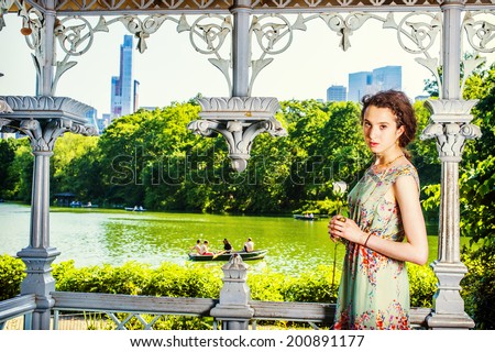 Young Lady Waiting for You. Dressing in sleeveless, long dress, a pretty teenage girl is standing inside a pavilion, hands holding a white rose, looking at you. people rolling boats in background.