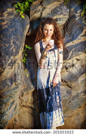 Dressing in long dress, chunky chain bracelet, arm cuff bracelet, a teenage college student with curly long hair is standing against rocks, hand holding a smart phone, smiling, reading, texting.
