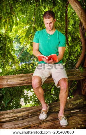 Young man reading outside. Wearing a green short sleeve Henley shirt, light yellow shorts, leather casual shoes, a young handsome college student is sitting on tree trunk in woods, reading a red book.