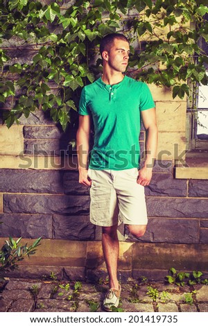 Young man waiting. Wearing a green short sleeve Henley shirt, light yellow shorts, leather casual shoes, bending a leg, a young guy is standing against the wall with green ivy leaves, looking around.