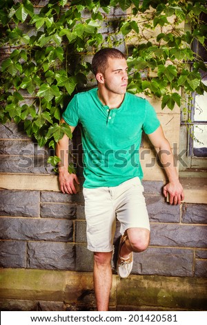 Young man waiting. Wearing a green short sleeve Henley shirt, light yellow shorts, bending a leg, a young guy is standing against the wall with green ivy leaves, looking around.