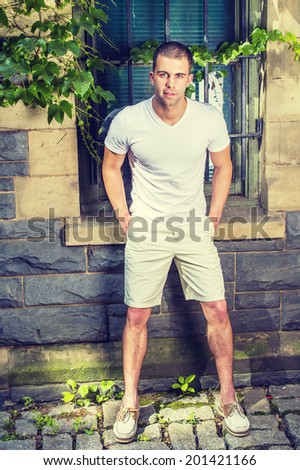 Young man. Wearing a white V neck T shirt, light yellow shorts,  leather shoes, hands in pockets, lowering back, a young guy is standing by a window with green ivy leaves, smiling, looking at you.