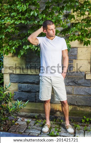 Young man thinking. Wearing a white V neck T shirt, light yellow shorts,  leather shoes, a hand scratching the back of head, a young guy is standing by a wall with green ivy leaves, waiting, thinking.