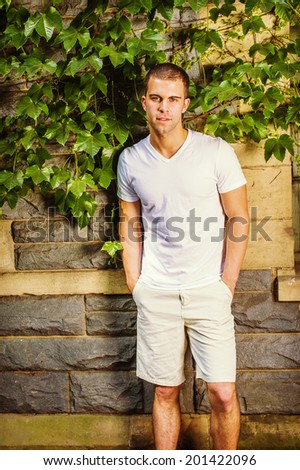 Portrait of young man. Wearing a white V neck T shirt, light yellow shorts, a young guy is standing against a wall with green ivy leaves, confidently looking at you.