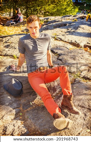 Man Waiting for You. Dressing in a gray long sleeves with roll-tab Henley shirt, red jeans, brown boot shoes,  a Fedora hat on side, a young sexy guy is sitting on rocks, relaxing, looking at you.