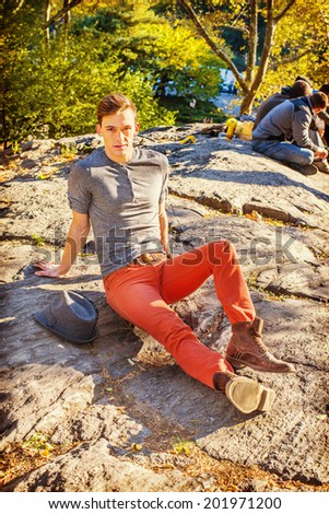 Man Waiting for You. Dressing in a gray long sleeves with roll-tab Henley shirt, red jeans, brown boot shoes,  a Fedora hat on side, a young sexy guy is sitting on rocks, relaxing, looking at you.