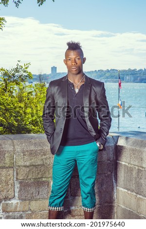 Man Traveling. Dressing in a black blazer, a black undershirt and green pants,  two hands putting in pockets, a young black guy with mohawk hair is standing by a river, relaxing. Urban Fashion