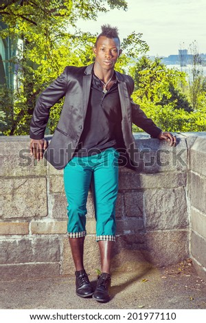 Man Relaxing Outside Dressing in a black blazer, a black undershirt, green pants, leather shoes, a young black guy with mohawk hair is standing by a rocky fence, relaxing. Urban Fashion.