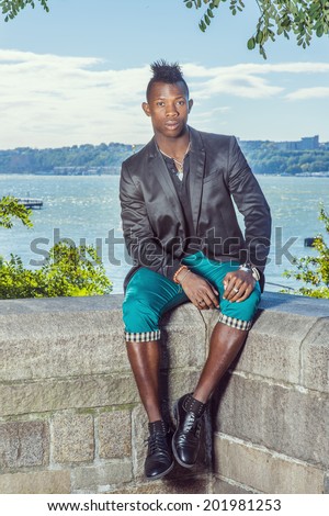 Man Relaxing Outside. Dressing in a black blazer,  green pants, leather shoes, a young black guy with mohawk hair is sitting on a rocky fence by a river, relaxing. Urban Fashion