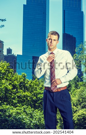 Portrait of Young Businessman. Wearing a white long sleeve shirt, black pants, a young professional guy is standing in the front of business district, tilting head, tying a tie, ready to work.