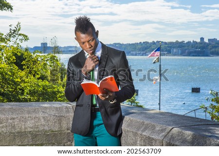 Young Man Reading Outside. Wearing a black blazer, green pants, a green tie, a hand touching chin, holding a red book, a young black guy with mohawk haircut is standing by a river, reading, thinking.