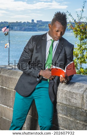 Young Man Reading Outside. Dressing in a white undershirt, a black blazer, green pants, a green tie, holding a red book,  a young black guy with mohawk haircut is standing by a river, reading outside.