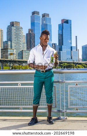 City Love. Wearing a white shirt,  green pants, leather shoes,  a young black guy with mohawk hair is standing in the front of buildings, looking at a white rose on his hands into deeply thinking.