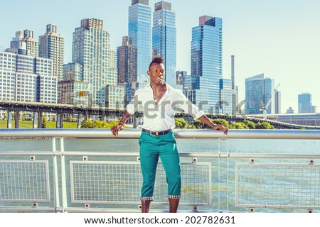 Urban Casual Fashion. Dressing in a white, long sleeve shirt, green pants,  a young black guy with mohawk haircut is standing in the front of business district, confidently looking forward.