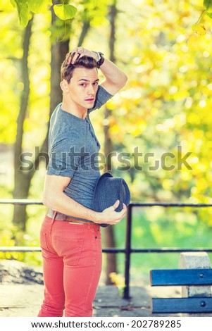 Man looking for You. Wearing a long sleeves with roll-tab shirt, red jeans, hand holding a woolen Fedora hat, touching hair, a young handsome guy is standing by golden autumn foliage, looking back.