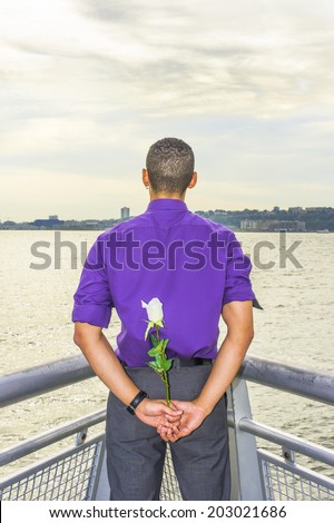 Young Man holding flower in back. Dressing in a purple shirt, gray pants, a young guy is standing by a river, facing water, holding a white rose behind his back,  waiting for you.