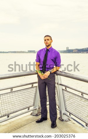 Young Man Waiting for You. Dressing in a purple shirt, gray pants, a black tie, holding a white flower, a young guy with beard, mustache is waiting for you by the river.