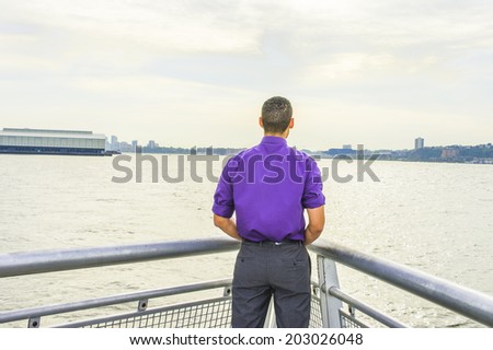 Young Man facing water in back view. Dressing in a purple shirt, gray pants, a young guy with shot curly hair is standing by a river, looking faraway, waiting for you.